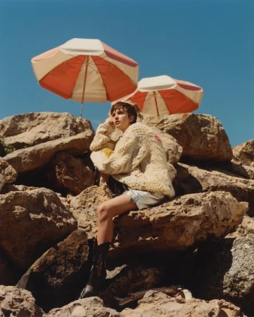 Young adult woman in a sweater and boots poses on rocks under two parasols against a blue sky on a sunny day in an outdoor setting.