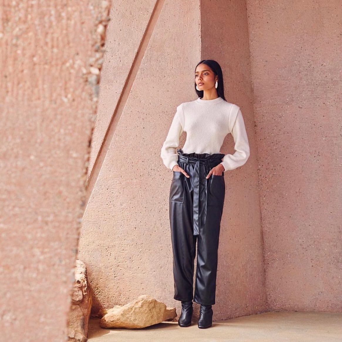 A brunette woman in an ivory sweater and black faux-leather paper-bag-waist pants stands in front of a textured peach-colored wall.