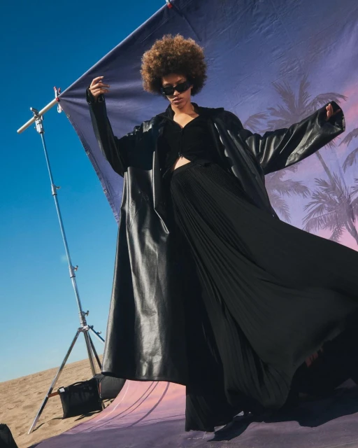 Woman in black coat and pleated pants poses against backdrop with palm trees under blue sky, photographed from a low angle.