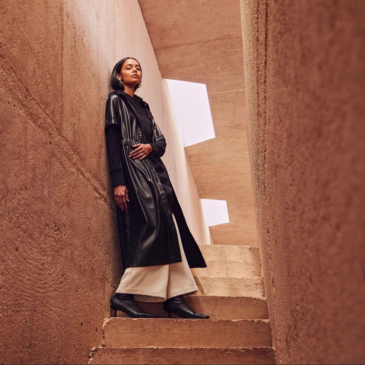 A woman in a black coat stands on concrete steps in an architectural setting. She is wearing a dark top, wide-leg pants, and black boots.