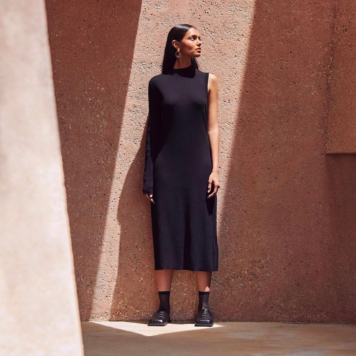 A full shot of a slender adult woman in a stylish black one-sleeve dress, black socks, and shoes, posing against a textured beige wall.