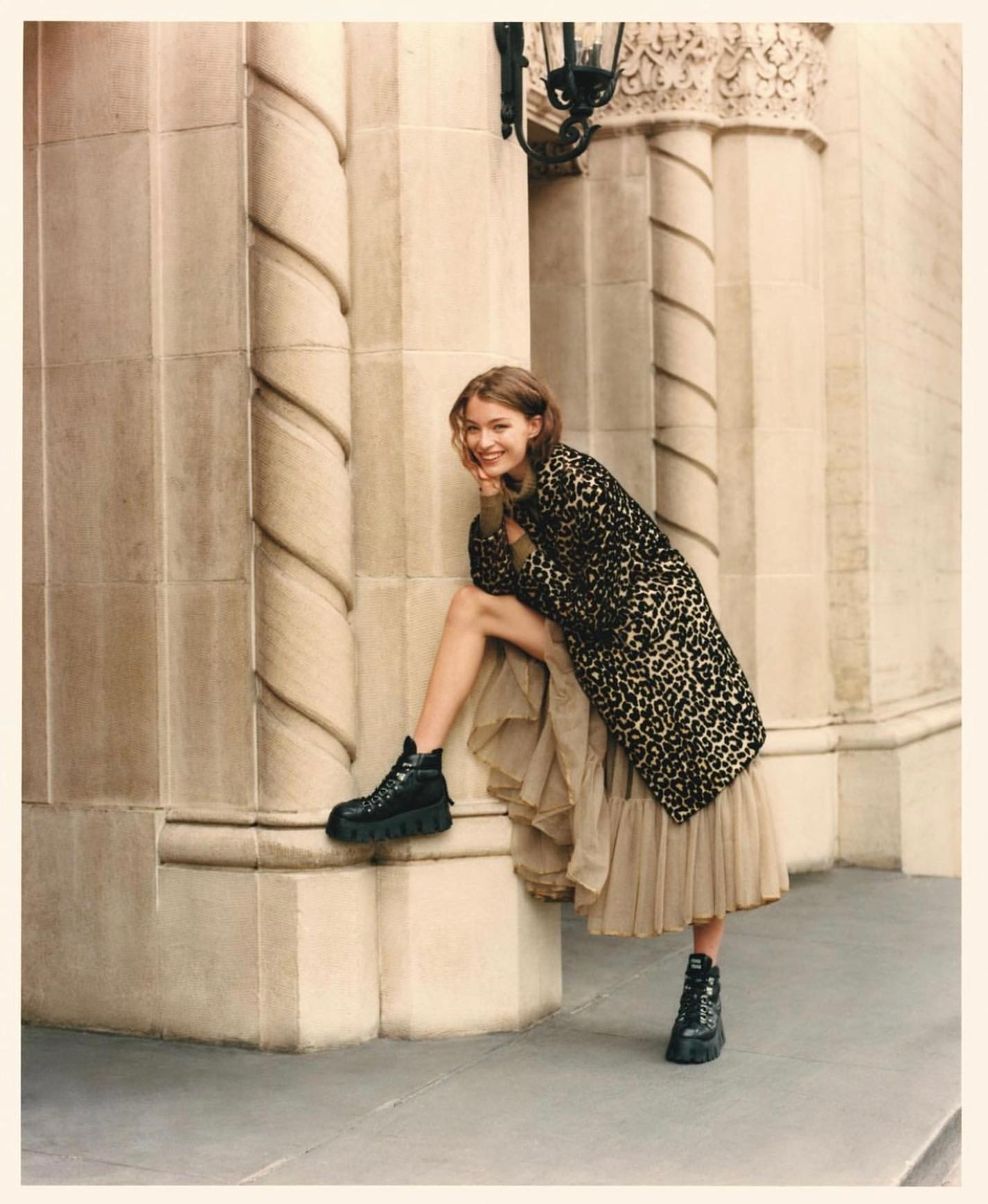 A woman in a leopard print coat and black platform boots poses in front of a stone building with carved columns and decorative elements.