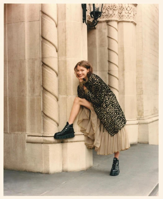 A woman in a leopard print coat and black platform boots poses in front of a stone building with carved columns and decorative elements.