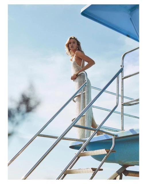 Blonde woman in a white dress stands on a lifeguard tower under blue sky. Fashion shot with natural light. Vertical composition.
