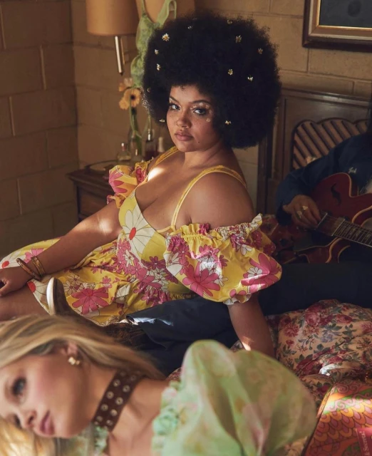 Two women are posing on a floral bedspread, one sitting with a large afro, the other laying down. A man plays guitar in background.