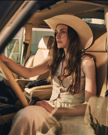 Woman in a beige vest and cowboy hat driving a car with tan interior.