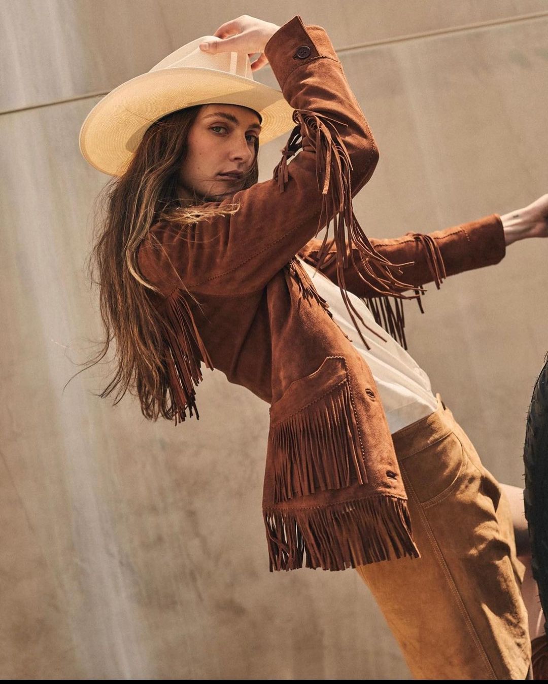 Brunette woman in fringed jacket and cowboy hat poses in front of a modern wall in a fashion editorial-style outdoor shot.