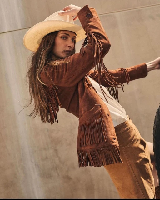Brunette woman in fringed jacket and cowboy hat poses in front of a modern wall in a fashion editorial-style outdoor shot.