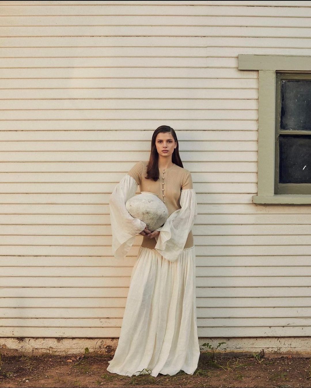 Woman in long skirt and knitted top poses with a round object outside a white wood-paneled house on a sunny day.