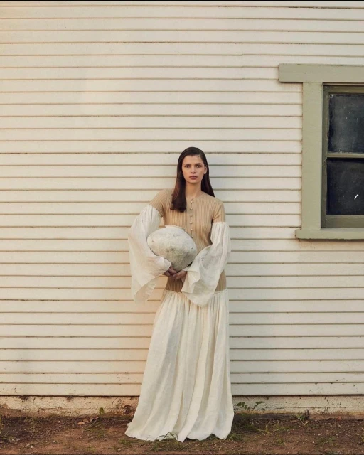 Woman in long skirt and knitted top poses with a round object outside a white wood-paneled house on a sunny day.