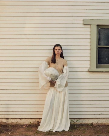 Woman in long skirt and knitted top poses with a round object outside a white wood-paneled house on a sunny day.