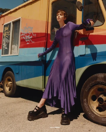 Model in a long purple dress and platform shoes leaning against a vibrant multi-colored food truck outdoors