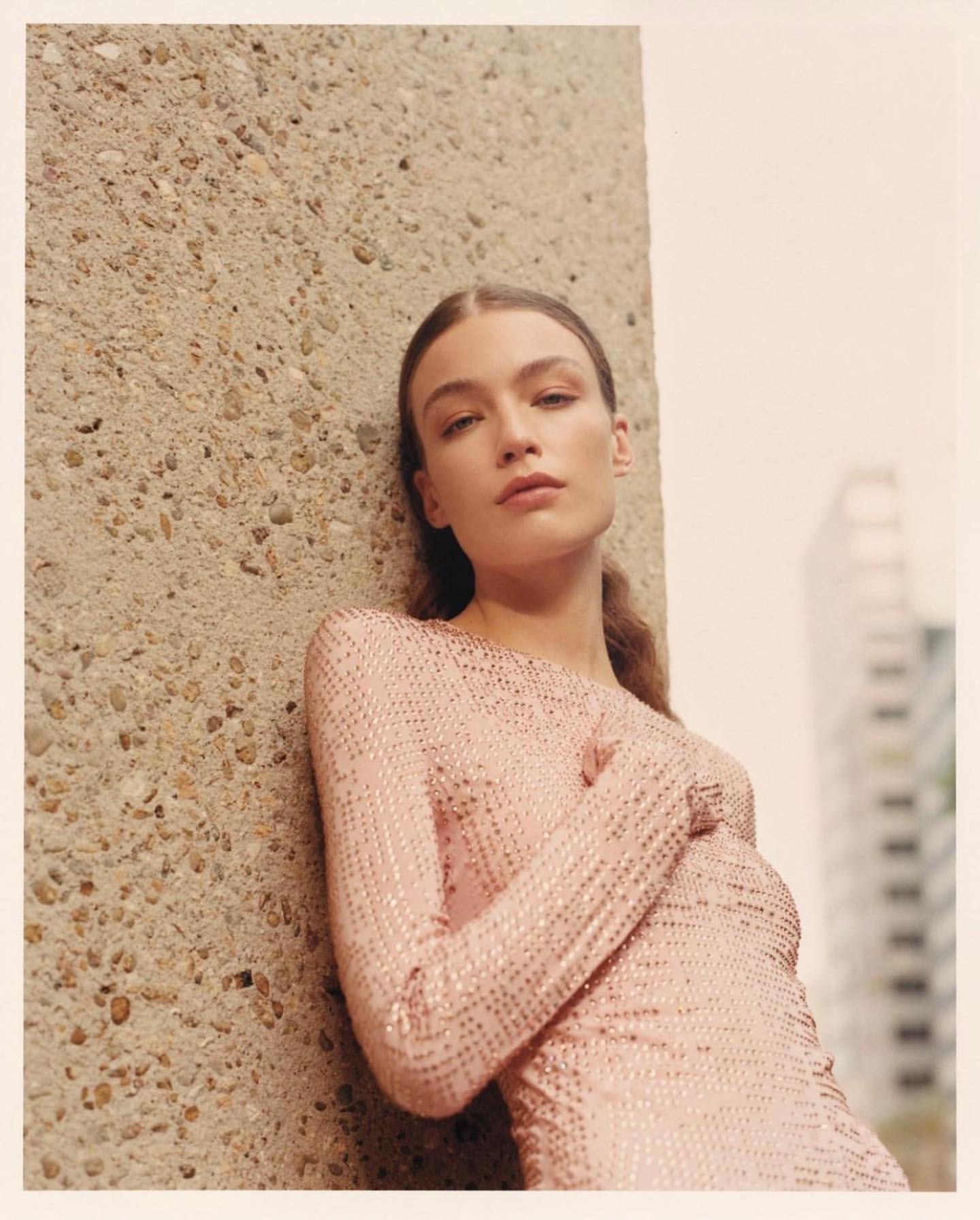 A young woman in a pale pink shimmering dress leans against a concrete pillar, posing in an outdoor urban setting with natural light.