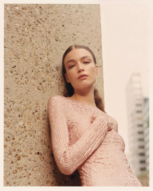 A young woman in a pale pink shimmering dress leans against a concrete pillar, posing in an outdoor urban setting with natural light.
