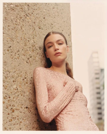 A young woman in a pale pink shimmering dress leans against a concrete pillar, posing in an outdoor urban setting with natural light.