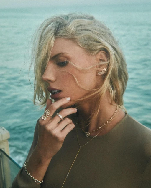 Close-up shot of a blonde woman in olive green wearing gold jewelry, including a necklace, rings, and earrings, posing in front of the ocean.