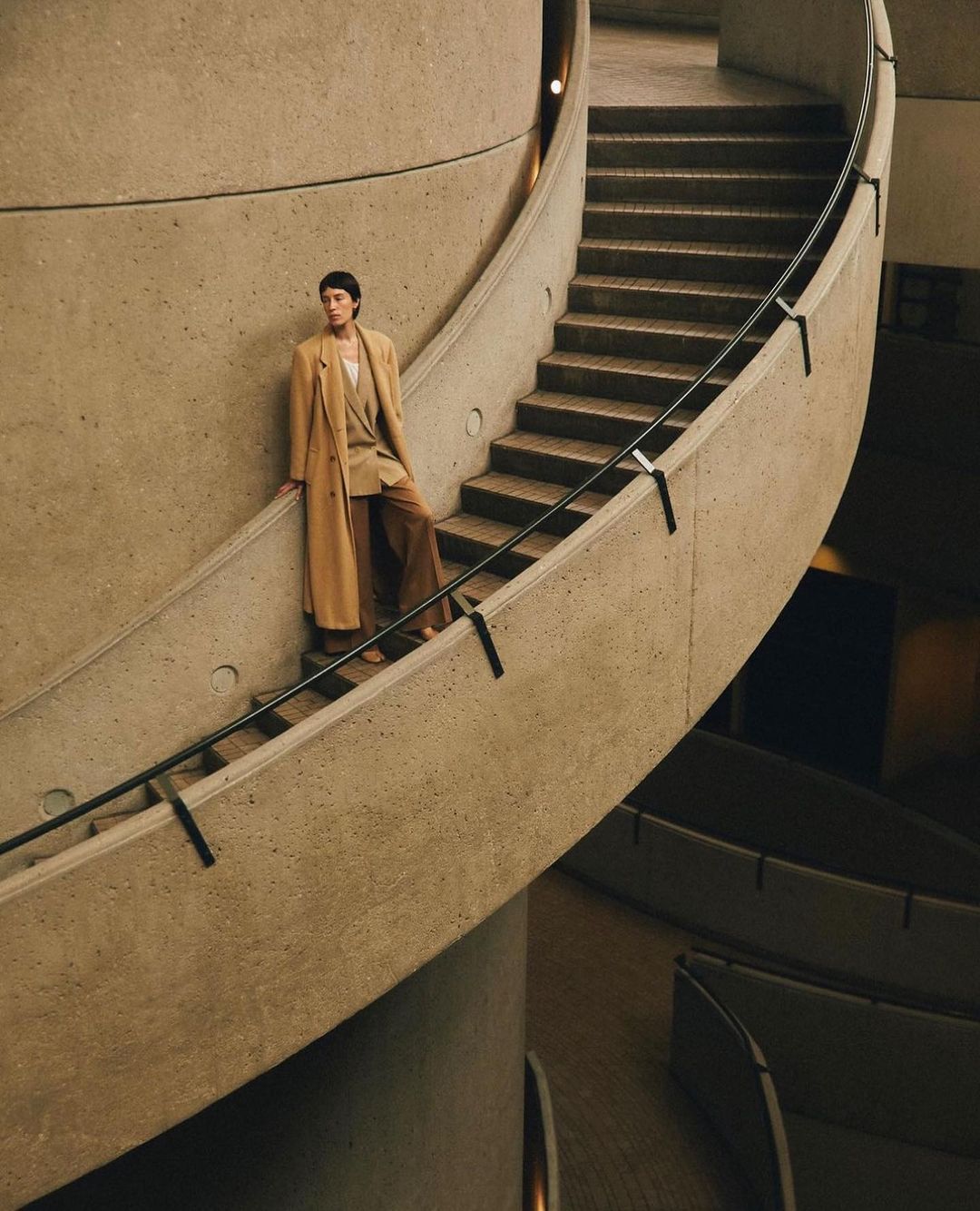 Model wearing a long beige coat standing on a curved concrete staircase by Justin Wilczynski.