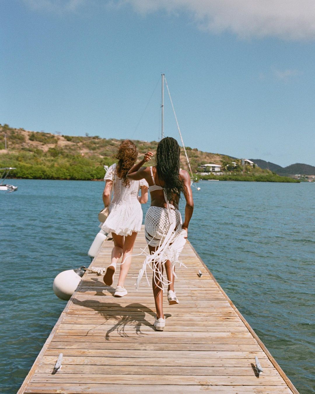 Two women in light dresses run on a wooden pier toward the ocean under a sunny sky, with boats and green hills in the background.