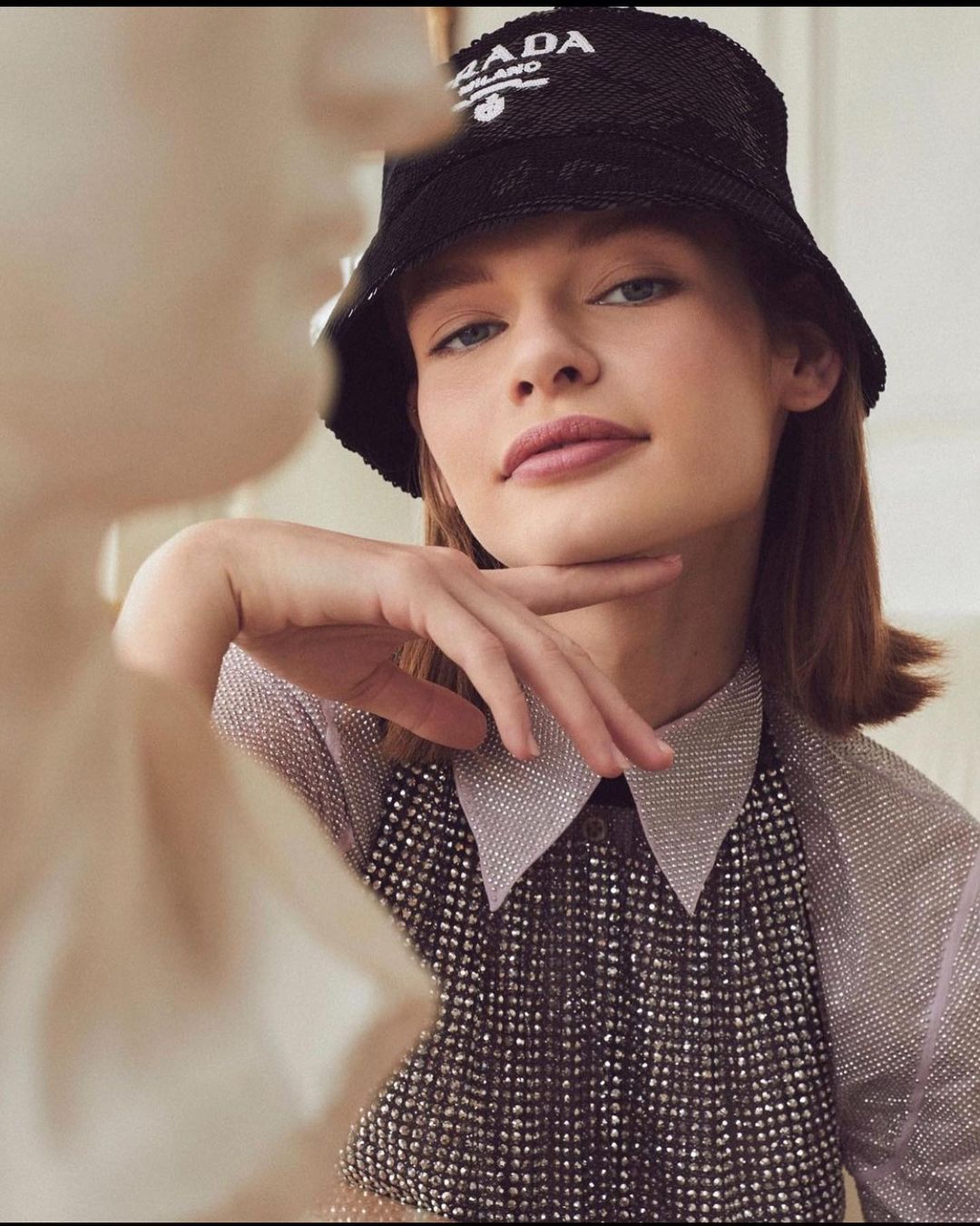 Close-up shot of a young woman wearing a Prada bucket hat and a sparkly top, posing with her hand to her chin in an indoor setting.