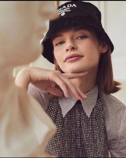 Close-up shot of a young woman wearing a Prada bucket hat and a sparkly top, posing with her hand to her chin in an indoor setting.