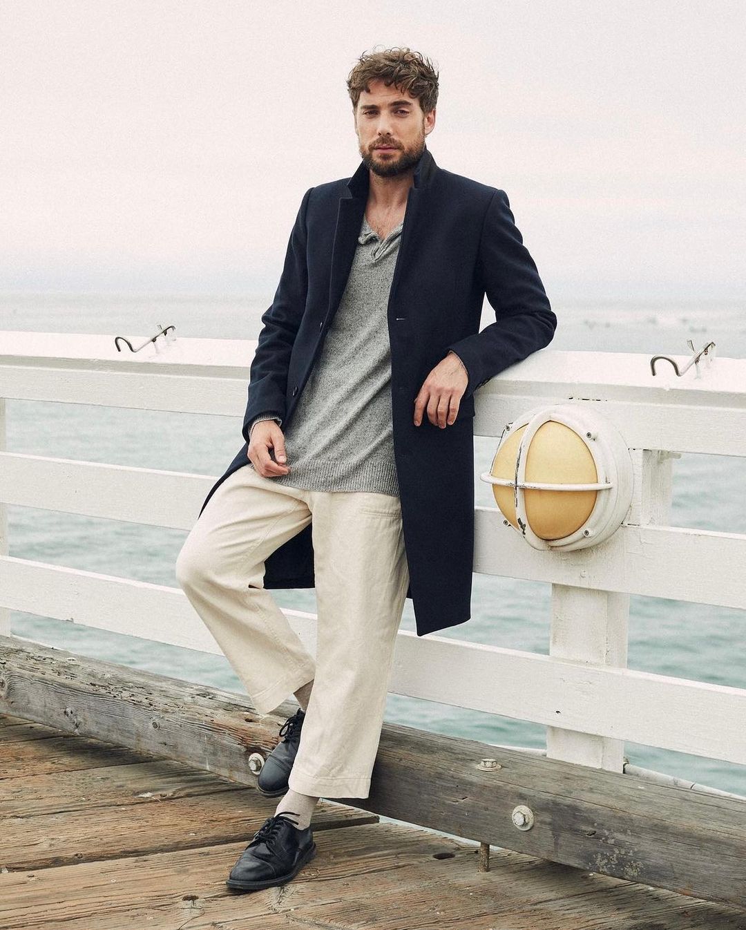 Adult male in a casual, fashionable outfit leans against a white pier railing with a calm ocean in the background on an overcast day.