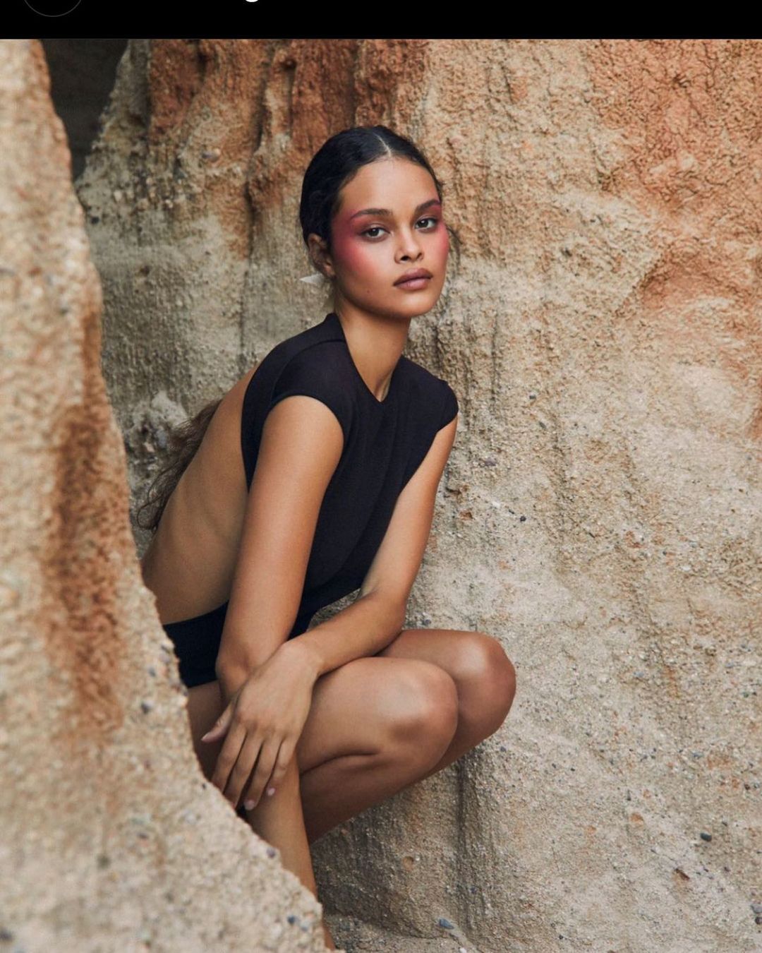 Attractive young woman in black sleeveless top poses in a crouching position near a light-colored rock formation, looking at the camera.