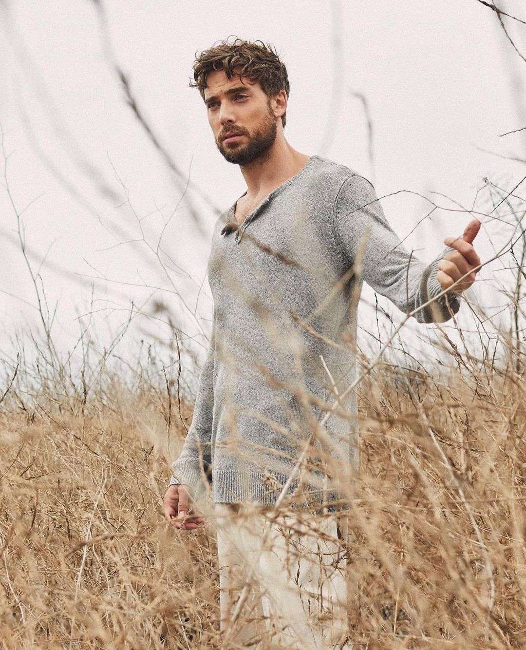 Man in a gray sweater and white pants stands among tall, dry grass in a neutral-toned, outdoor environment with soft, diffused lighting.