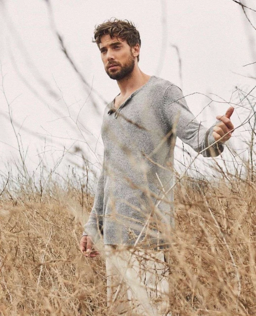 Man in a gray sweater and white pants stands among tall, dry grass in a neutral-toned, outdoor environment with soft, diffused lighting.