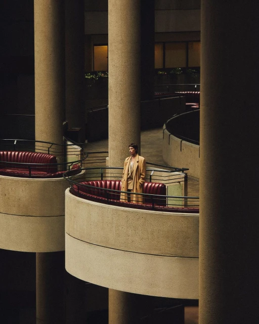 Adult stands on balcony with terracotta sofas inside brutalist building with beige columns and railings in a medium, eye-level shot.