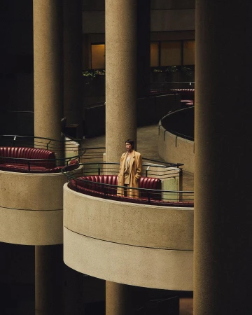 Adult stands on balcony with terracotta sofas inside brutalist building with beige columns and railings in a medium, eye-level shot.