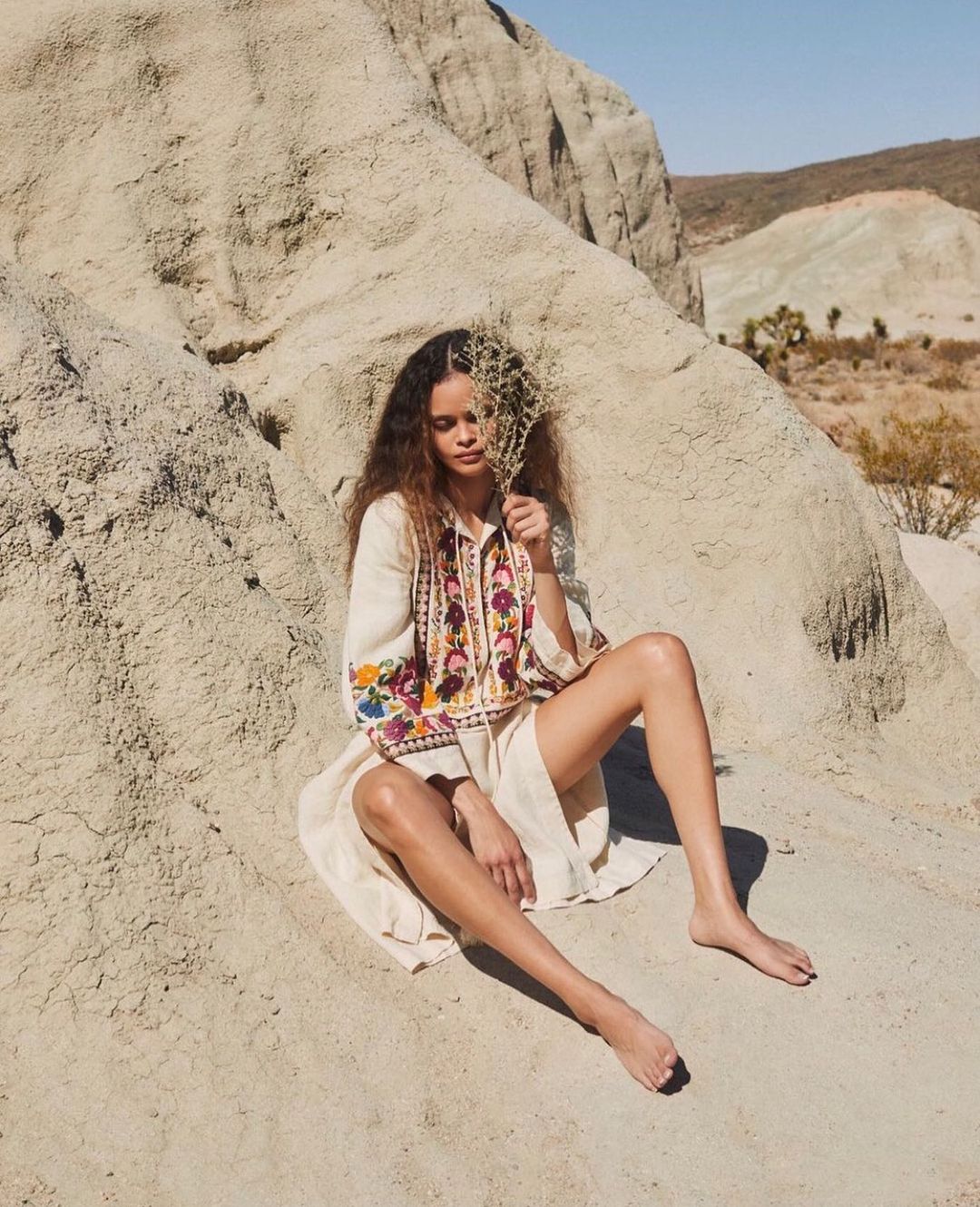 Adult female poses in a desert setting wearing an embroidered tunic and skirt, sitting barefoot on a rock formation on a clear day.