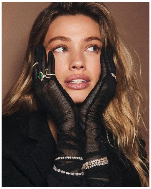 Portrait of a fair-skinned adult female with long blonde hair, wearing black gloves and jewelry, posing against a neutral background.