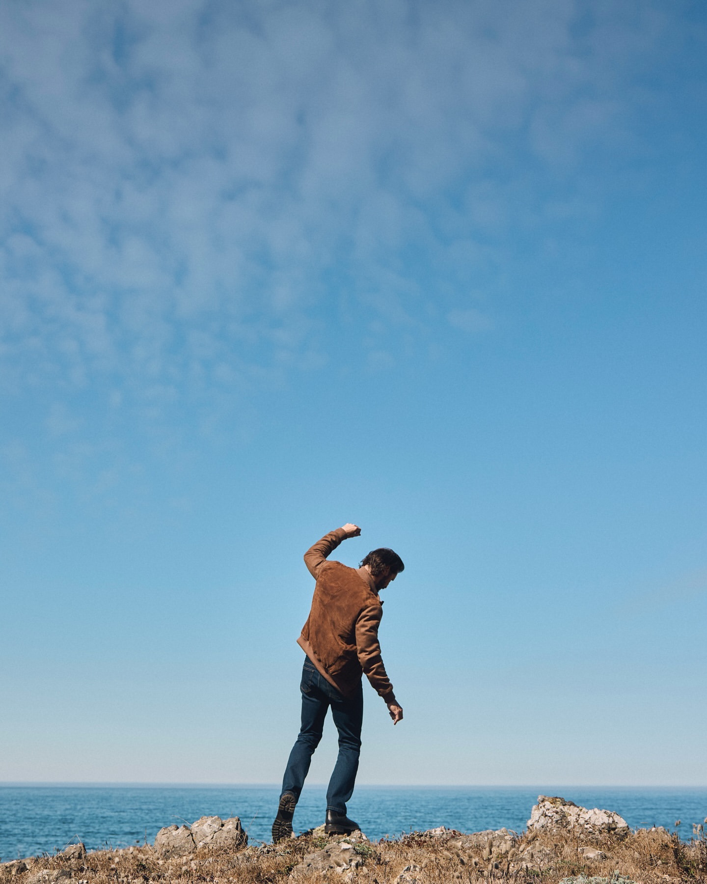 A man in a brown jacket stands on a rocky cliff against a blue sky and ocean backdrop, arm raised near the coastline. 