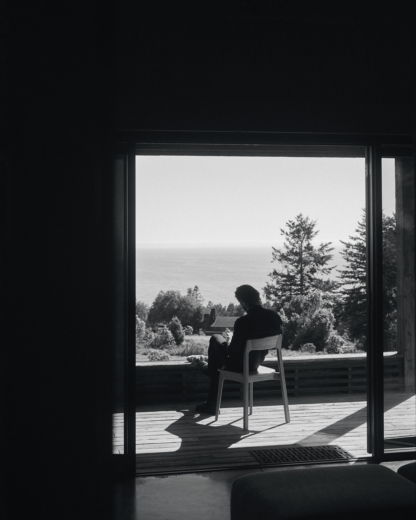 Monochrome portrait of a seated man, back to camera, gazing through sliding glass doors at a distant sea and trees, outdoors.