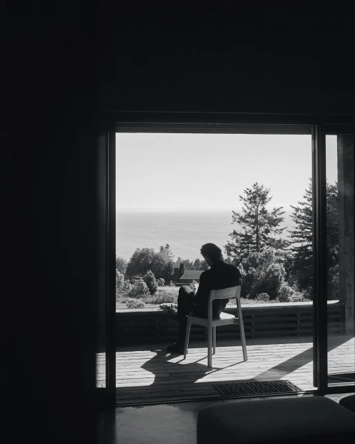 Monochrome portrait of a seated man, back to camera, gazing through sliding glass doors at a distant sea and trees, outdoors.
