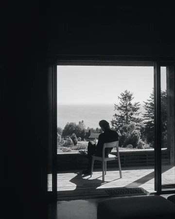 Monochrome portrait of a seated man, back to camera, gazing through sliding glass doors at a distant sea and trees, outdoors.