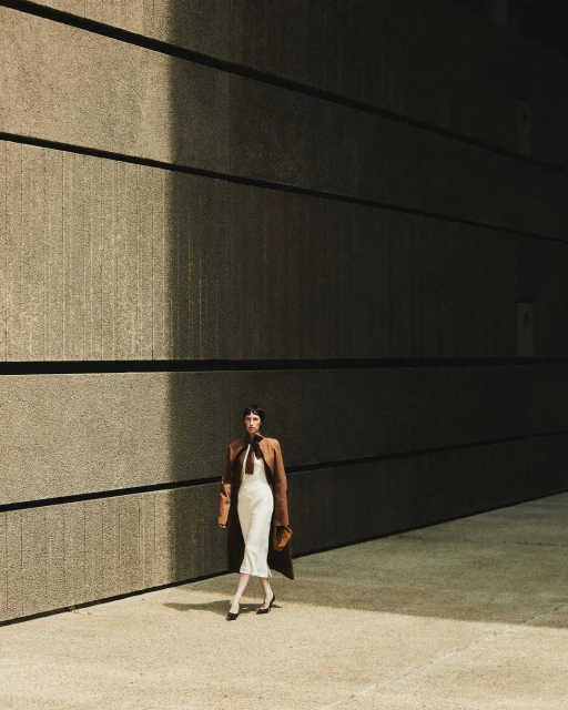 Woman in a brown coat and white dress walking in an urban setting with neutral concrete walls and ground, creating a minimalist fashion shot.