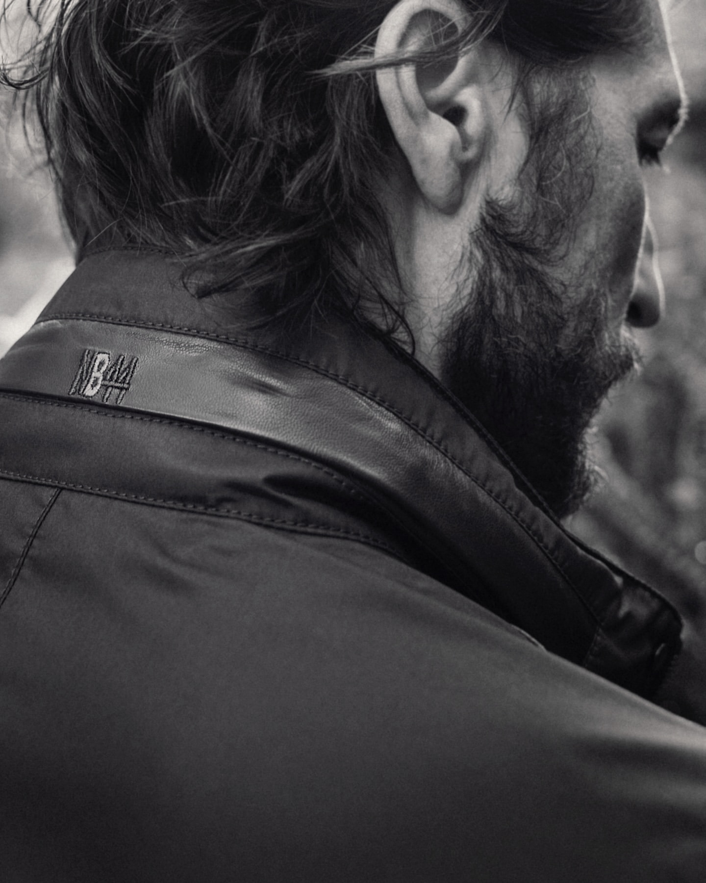 Black and white close-up of an adult male's head and shoulders, showcasing a dark jacket with subtle logo detail at the collar.
