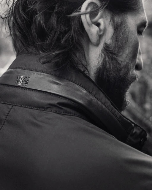 Black and white close-up of an adult male's head and shoulders, showcasing a dark jacket with subtle logo detail at the collar.