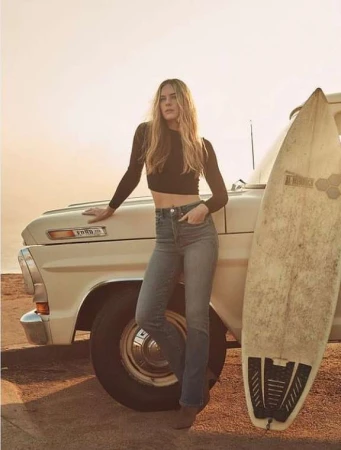 Woman with long blonde hair leaning against a vintage pickup truck next to a surfboard at sunset