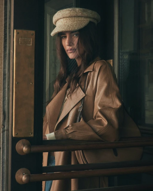 Portrait of a young adult female with long brown hair, wearing a trench coat and cap, posing in front of a dark door.