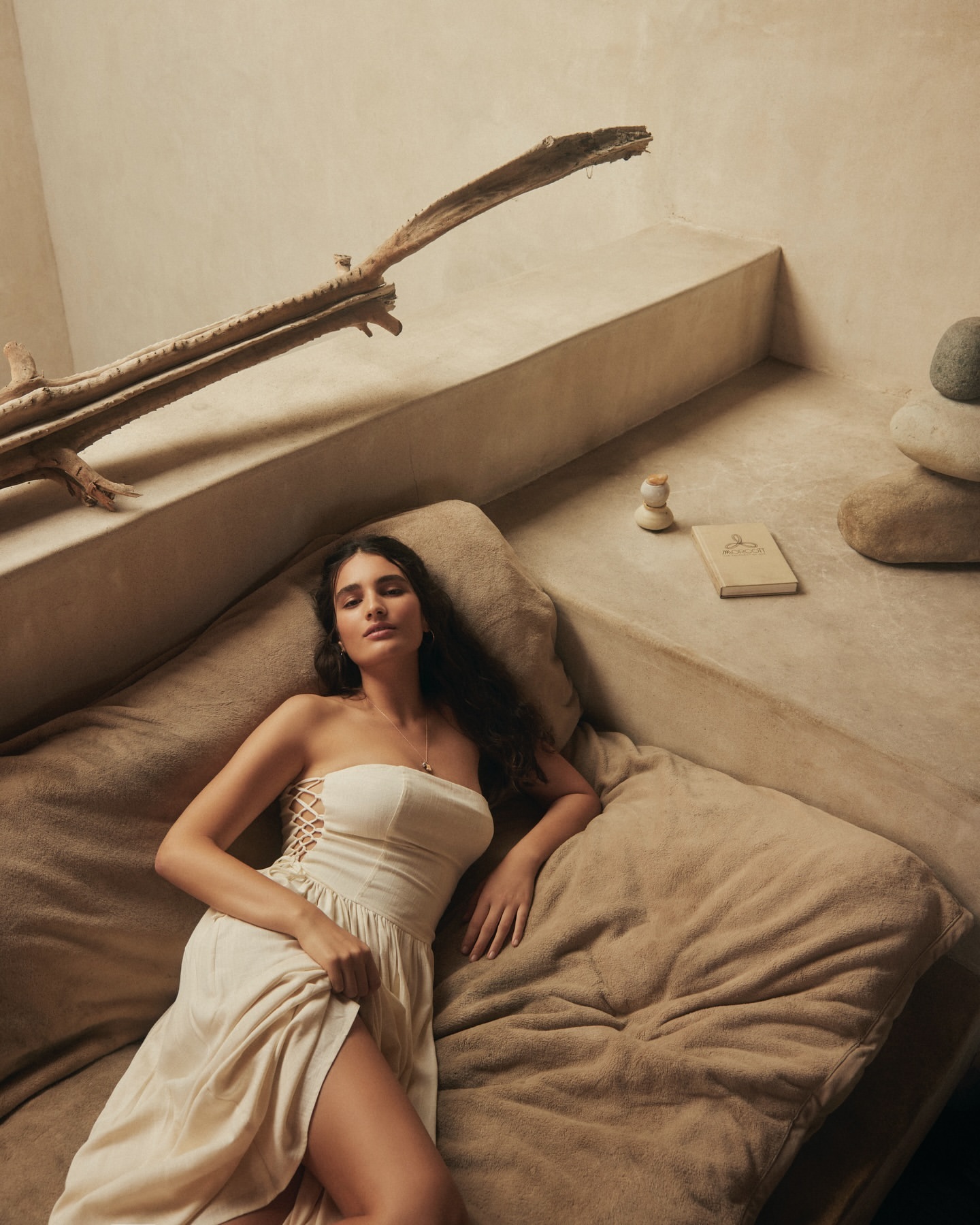 A woman with long dark hair is lying on a bed wearing an off-white dress in an indoor environment with soft lighting and natural textures.