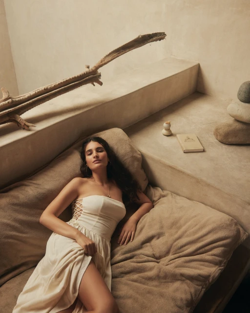 A woman with long dark hair is lying on a bed wearing an off-white dress in an indoor environment with soft lighting and natural textures.