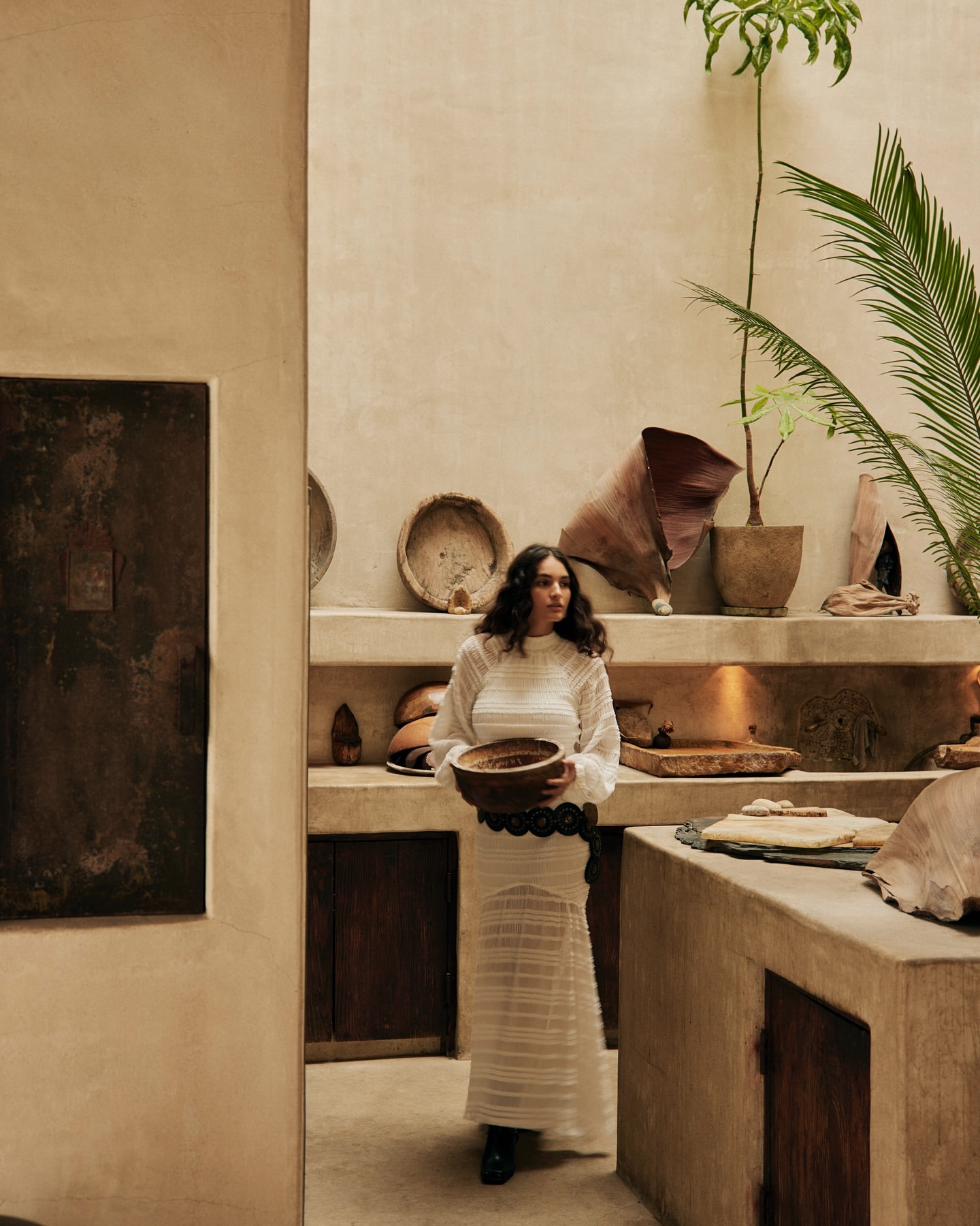 A woman in a white dress holds a bowl in a bright, neutral indoor setting with pots, woven baskets, and various clay objects on shelves.