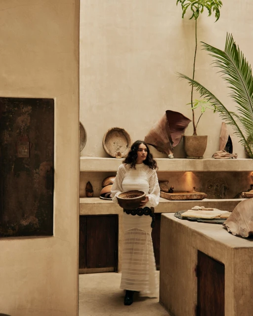 A woman in a white dress holds a bowl in a bright, neutral indoor setting with pots, woven baskets, and various clay objects on shelves.