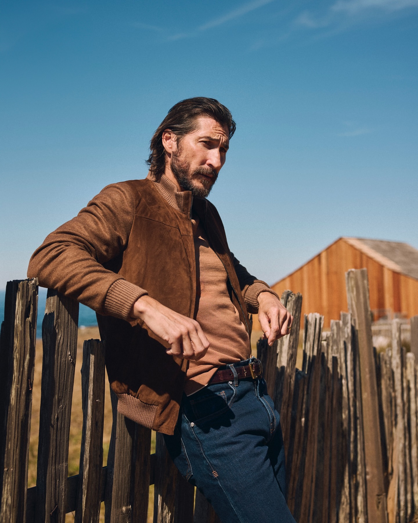 Man in a brown jacket leans against a wooden fence outdoors on a sunny day, with a building visible in the background.