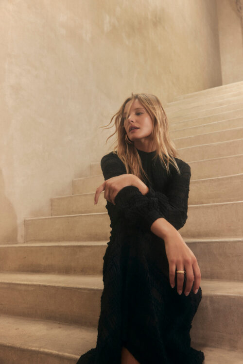 Blonde woman in a black dress sits on light-colored stairs with a textured wall. Soft lighting highlights the dress's texture.