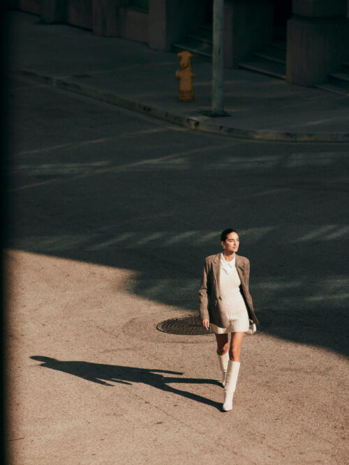 A young adult woman in a tan blazer and white shorts walks on a city street with cast shadows. She is wearing white boots.