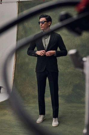 Man wearing sunglasses, black suit jacket, trousers, and white sneakers posing in front of a green textured backdrop.