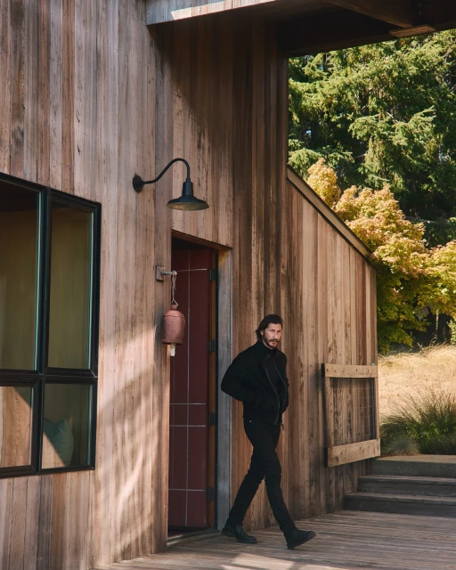 Adult male in black outerwear walks on a wooden porch outside a rustic wood building with greenery visible in the background on a sunny day.